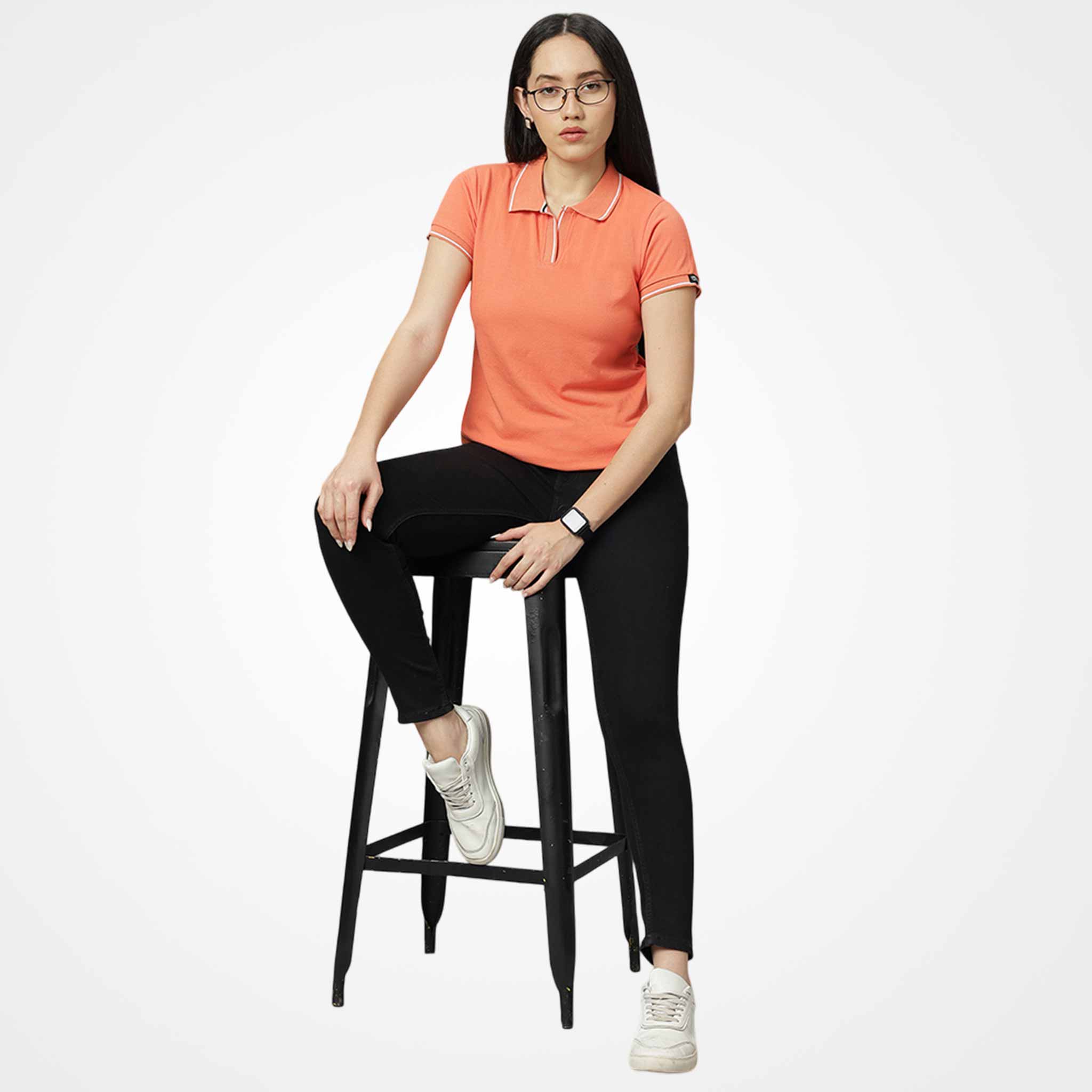 WYO Woman sitting on a black stool wearing an orange polo shirt and black pants against a white background