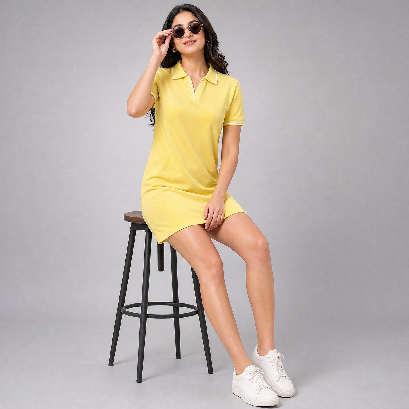 A woman wearing a WYO banana yellow short-sleeve polo dress sits on a black metal stool against a neutral gray studio background. She is wearing white sneakers and sunglasses.