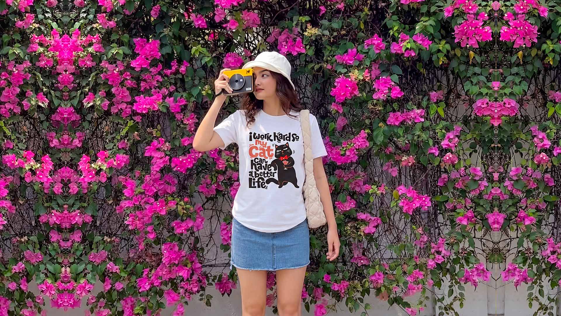 Women wearing a WYO White T-shirt and taking a photo with a camera against a vibrant pink flower wall 