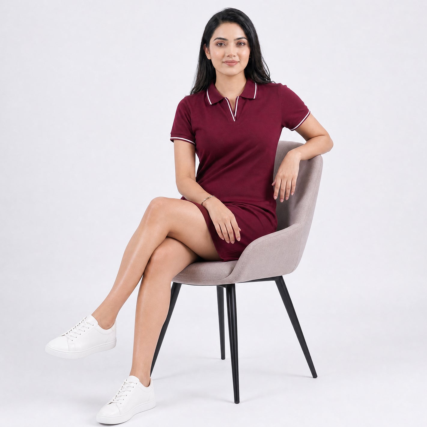A woman sits on a light gray upholstered chair wearing a WYO short-sleeve polo dress maroon with white piping on the collar and sleeve cuffs, paired with white sneakers, against a plain light gray studio background.