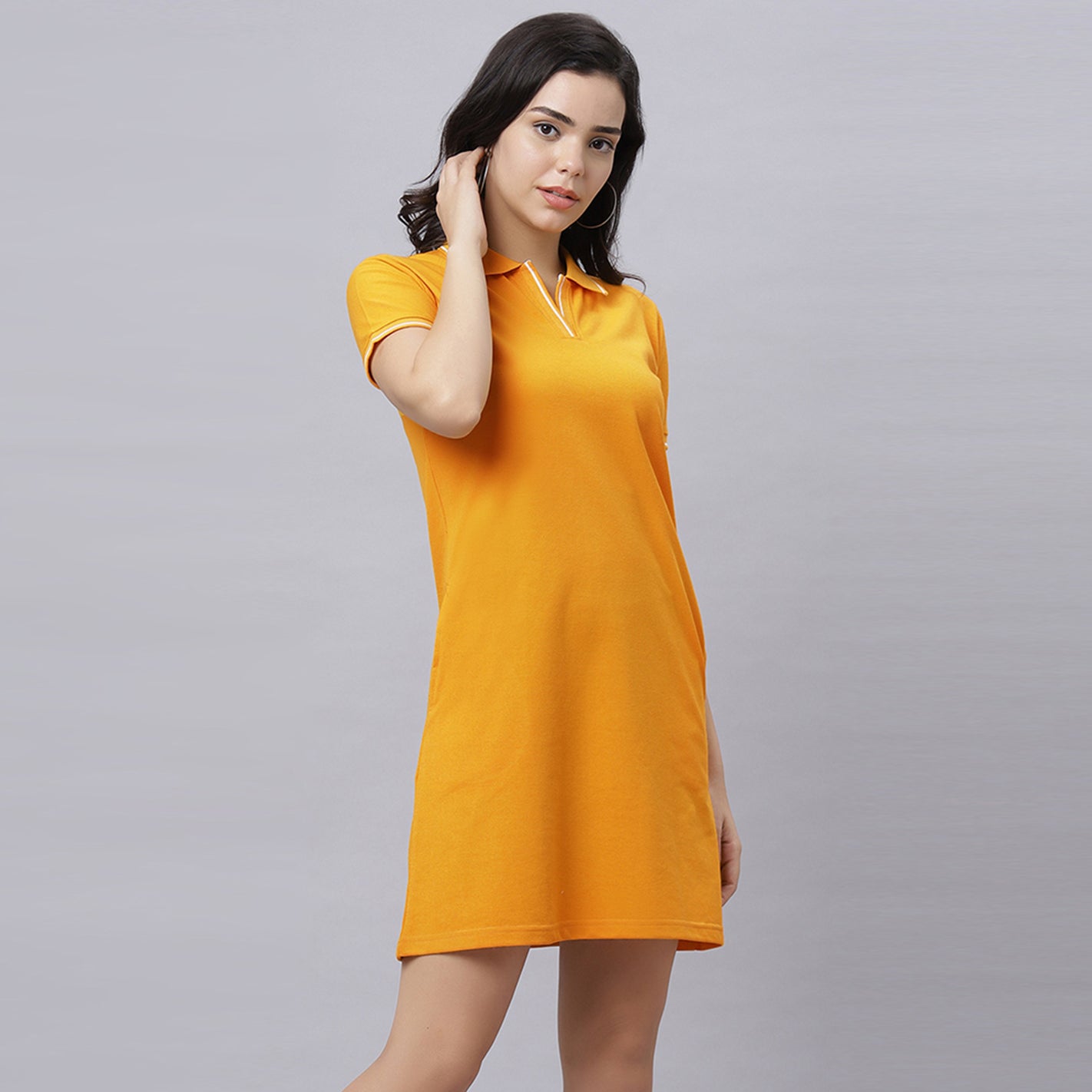A woman modeling a WYO short-sleeve polo shirt dress in a vibrant Mustard color, featuring a V-neckline with thin white piping details. She is posed against a solid light gray studio background.