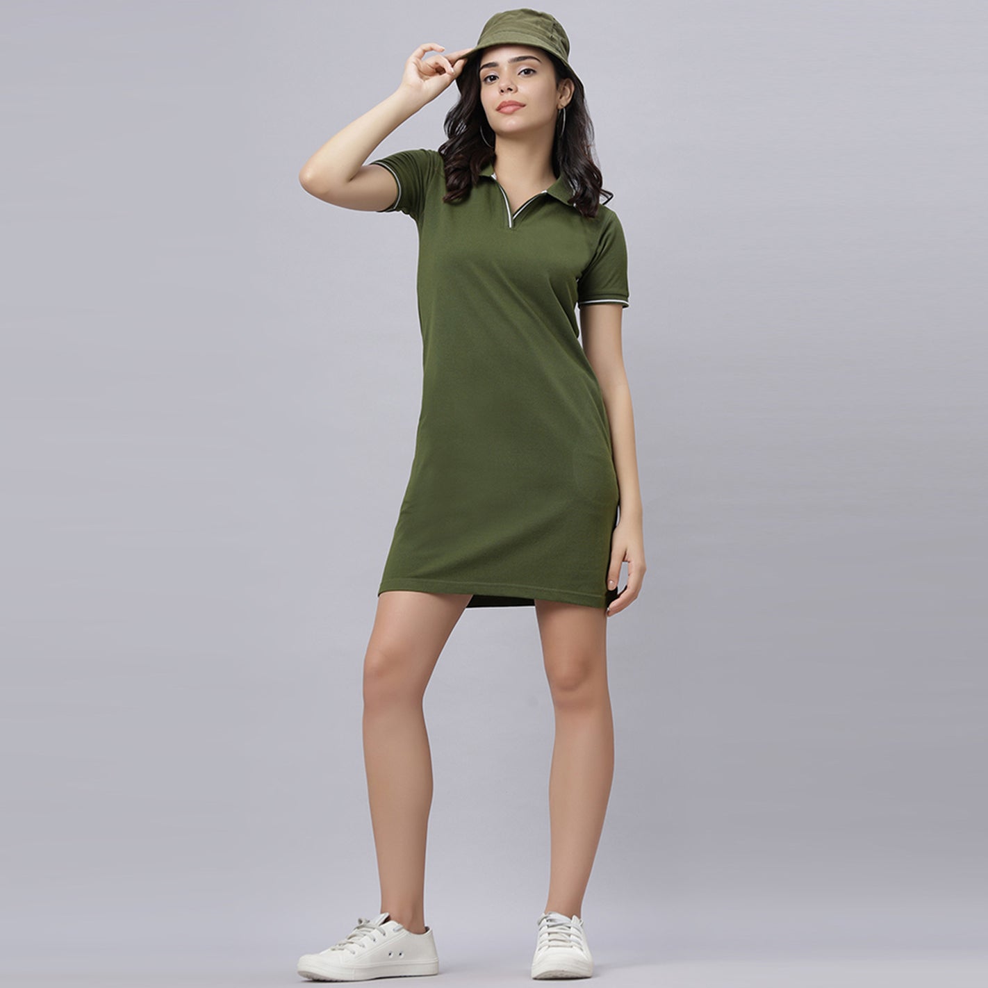 A woman modeling an WYO olive short-sleeve polo dress with white piping trim, paired with a matching olive green bucket hat and white sneakers, against a solid gray studio background.