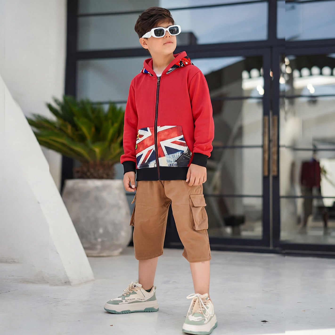 Child wearing a WYO red hoodie with a Union Jack design, brown cargo shorts, and white sneakers outdoors.