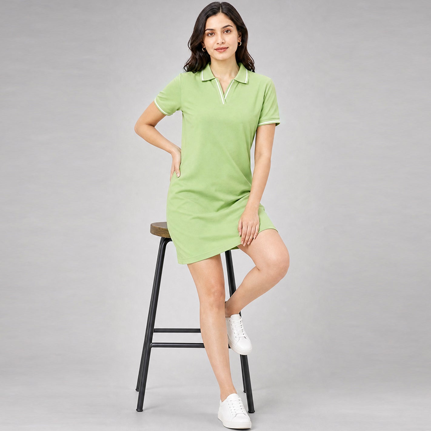 A woman models a WYO saga green short-sleeve polo dress while sitting on a black metal stool against a solid gray studio background.